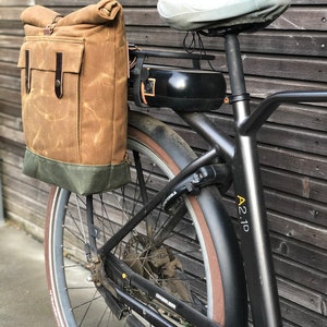 May include: A black electric bicycle with a brown canvas bag attached to the rear rack. The bicycle has a brown leather seat and a black battery pack. The bicycle has a black fender with the text "A21D" printed on it.