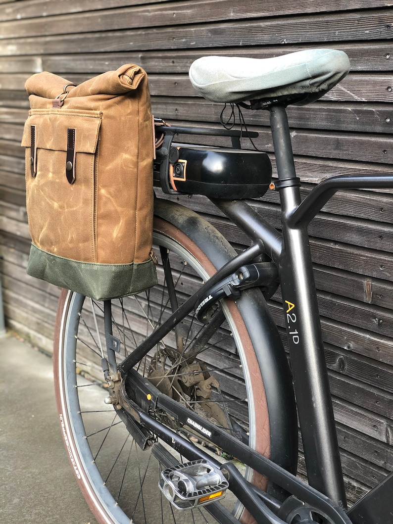 May include: A black electric bicycle with a brown canvas bag attached to the rear rack. The bicycle has a brown leather seat and a black battery pack. The bicycle has a black fender with the text "A21D" printed on it.