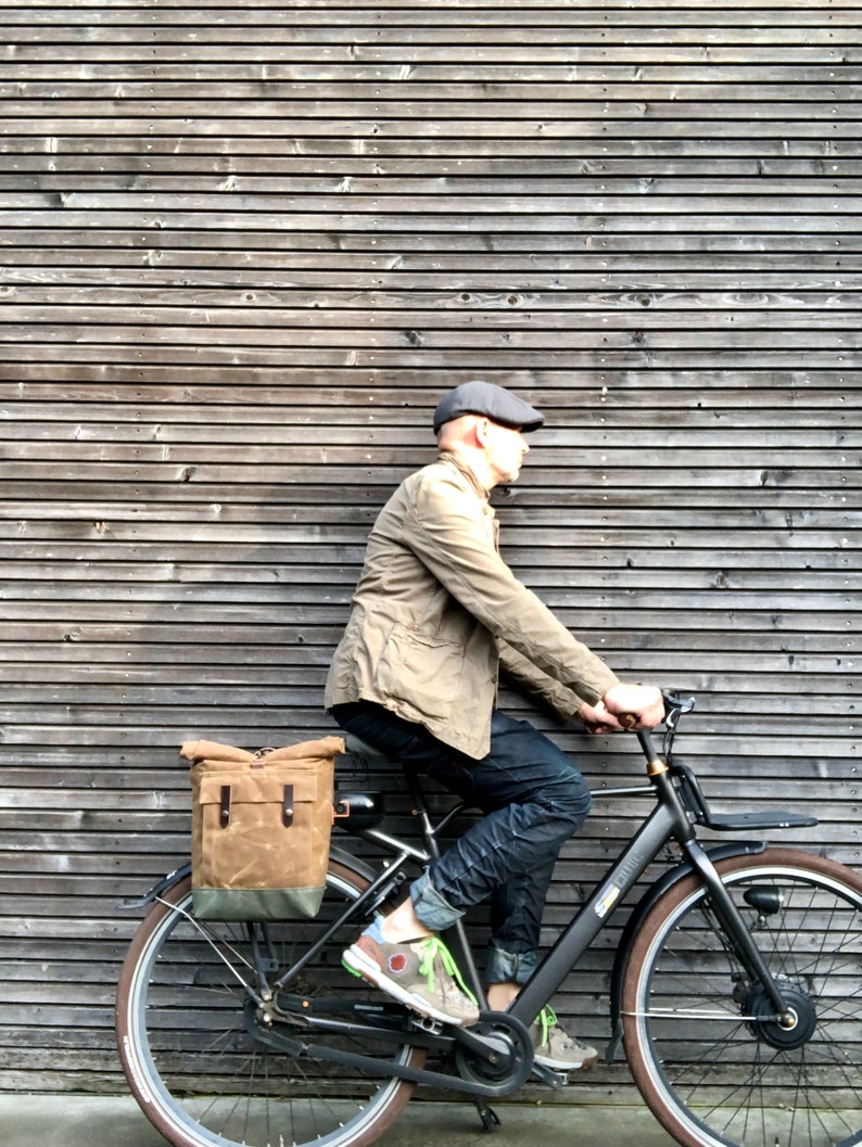 May include: A person riding a black electric bicycle with a brown canvas pannier bag attached to the rear rack. The bicycle has a black frame, black wheels, and a black seat.