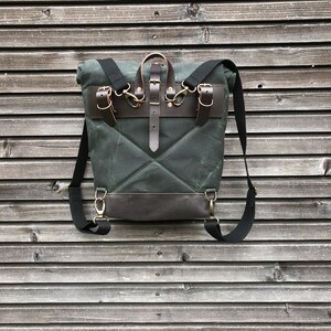 May include: A dark green canvas backpack with brown leather straps and buckles. The backpack has a flap closure and a small pocket on the front.