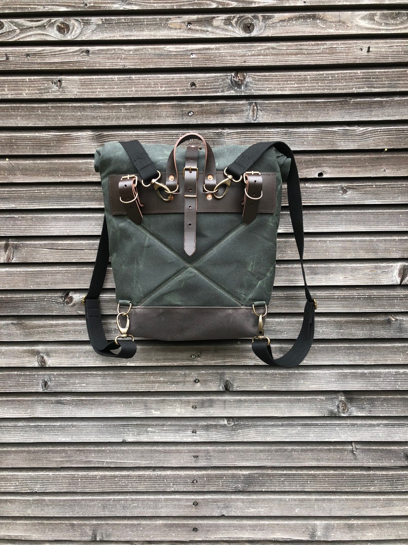 May include: A dark green canvas backpack with brown leather straps and buckles. The backpack has a flap closure and a small pocket on the front.