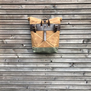 May include: A tan canvas backpack with brown leather straps and buckles. The backpack has a flap closure and a green canvas lining. The backpack is hanging on a wooden wall.