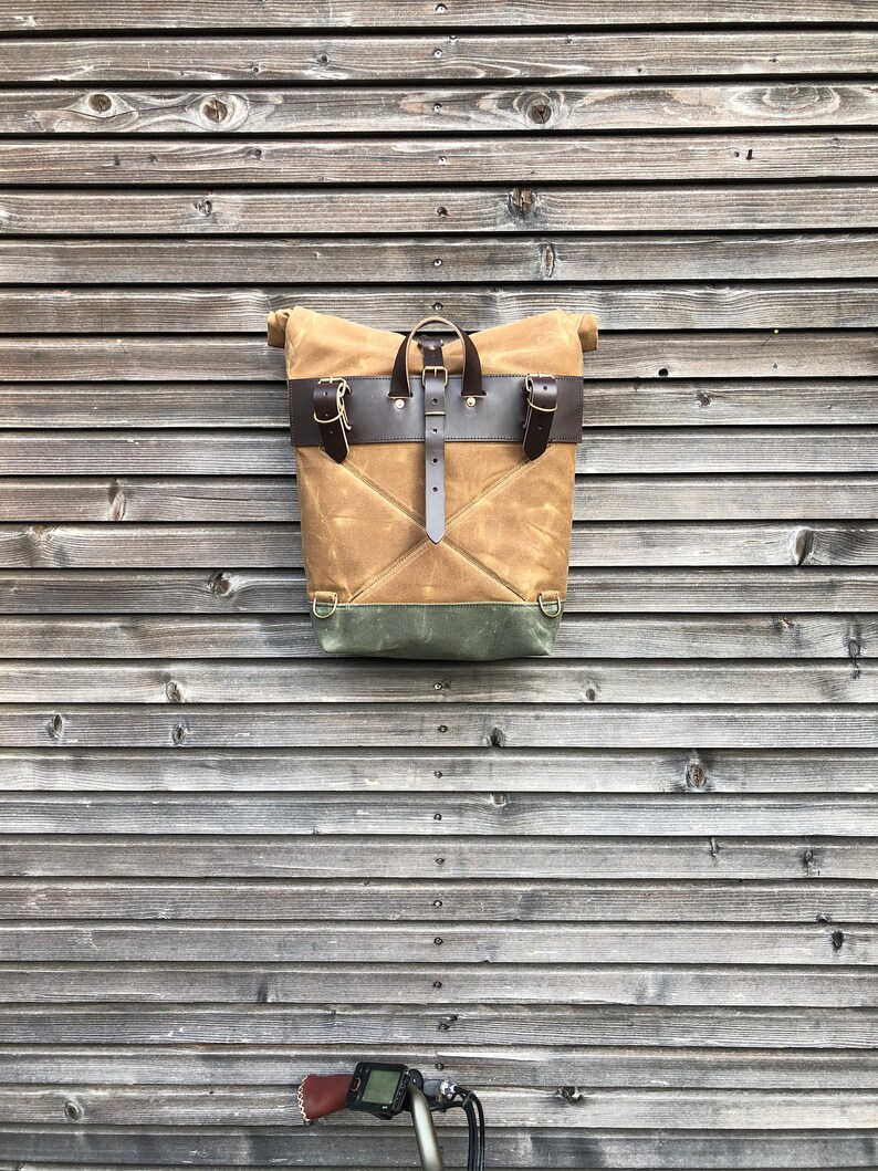 May include: A tan canvas backpack with brown leather straps and buckles. The backpack has a flap closure and a green canvas lining. The backpack is hanging on a wooden wall.