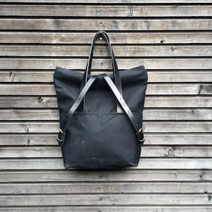 May include: A black canvas tote bag with leather straps and gold buckles. The bag is hanging on a wooden wall.