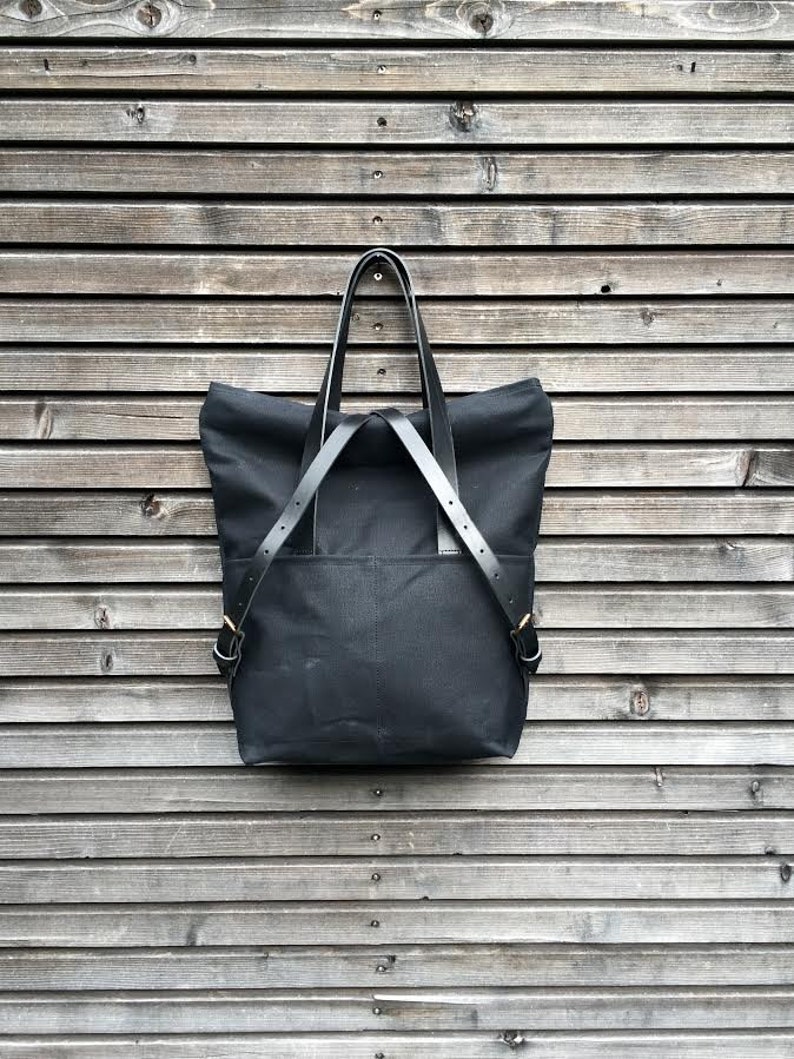 May include: A black canvas tote bag with leather straps and gold buckles. The bag is hanging on a wooden wall.