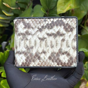 May include: A bi-fold wallet with a snakeskin pattern in shades of white, gray, and brown. The wallet has a black border and the text "Erios Leather" is visible. The wallet is held by a gloved hand.