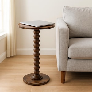 May include: A dark brown, round side table with a stacked ball design on the central support. A silver laptop rests on the top surface. The table is approximately 24 inches tall and is next to a light gray sofa.