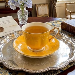 May include: A bright yellow porcelain teacup and matching saucer filled with tea, resting on a round, ornate silver-plated serving tray with decorative handles and an intricate embossed floral pattern along the scalloped rim.