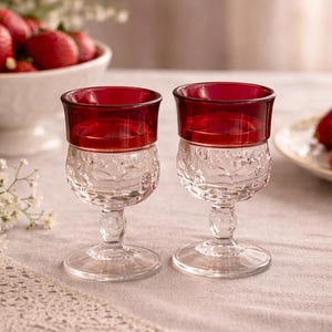 May include: Two clear glass goblets with ruby red rims. The lower portion of the glasses features an embossed floral design. The goblets are displayed on a light-colored surface, with a bowl of strawberries and flowers in the background.