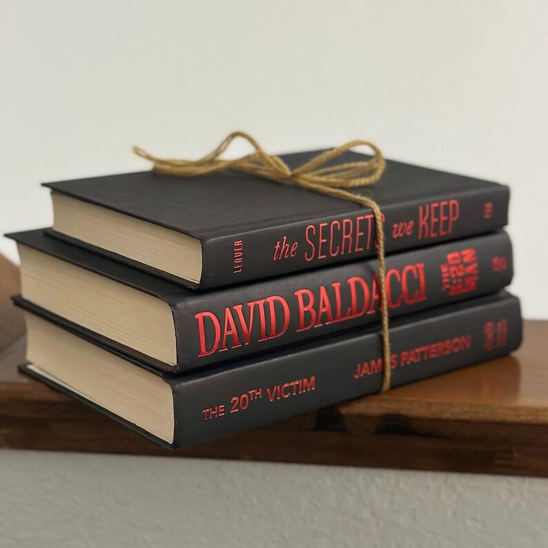 May include: Three hardcover books with black covers and red lettering are stacked and tied with twine. The titles include "The Secret We Keep," "David Baldacci," and "The 20th Victim."