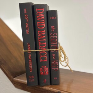 May include: Three hardcover books with black covers and red lettering are tied together with twine. The books are titled "The 20th Victim" by James Patterson, "David Baldacci The 6:20 Man", and "The Secrets We Keep". The books are on a wooden shelf.