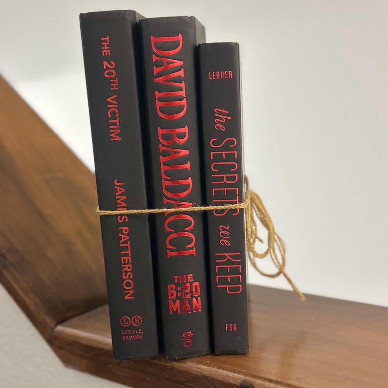 May include: Three hardcover books with black covers and red lettering are tied together with twine. The books are titled "The 20th Victim" by James Patterson, "David Baldacci The 6:20 Man", and "The Secrets We Keep". The books are on a wooden shelf.