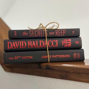 May include: Three hardcover books stacked on a wooden surface, bound together with twine. The books are black with red lettering. Titles include "The Secrets We Keep", "David Baldacci", and "The 20th Victim".