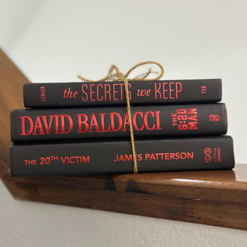 May include: Three hardcover books stacked on a wooden surface, bound together with twine. The books are black with red lettering. Titles include "The Secrets We Keep", "David Baldacci", and "The 20th Victim".