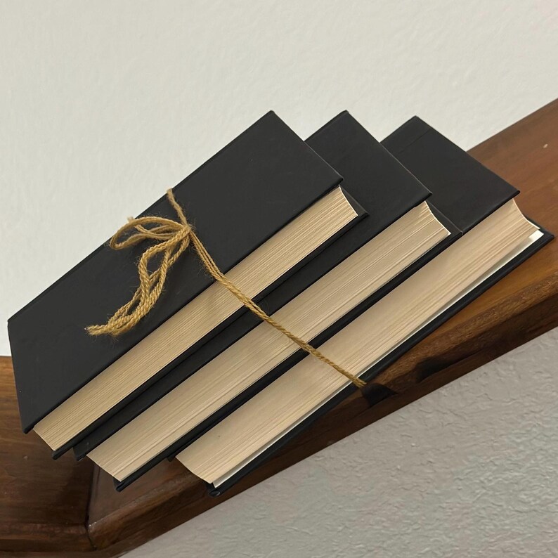 May include: Four black hardcover books with cream-colored pages are stacked on a wooden surface. A natural twine is wrapped around the books, tied in a bow. The books are angled, creating a visually appealing arrangement.