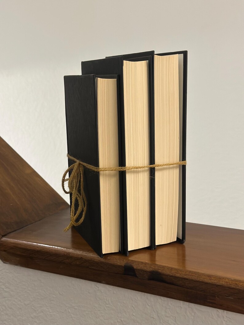 May include: Three hardcover books with black covers and cream-colored pages are tied together with twine. The books are displayed on a dark wooden shelf against a white wall. The spines of the books are facing outward.