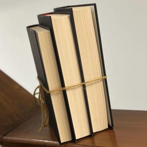 May include: Three hardcover books with black covers and cream-colored pages are tied together with twine. The books are stacked at an angle, with the spines facing the viewer. The edges of the pages are visible, and the binding is black.