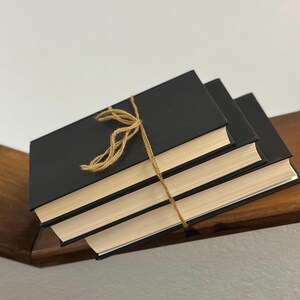 May include: Three black hardcover books tied together with twine. The books have cream-colored pages and are stacked on a wooden shelf. The twine is tied in a decorative bow.