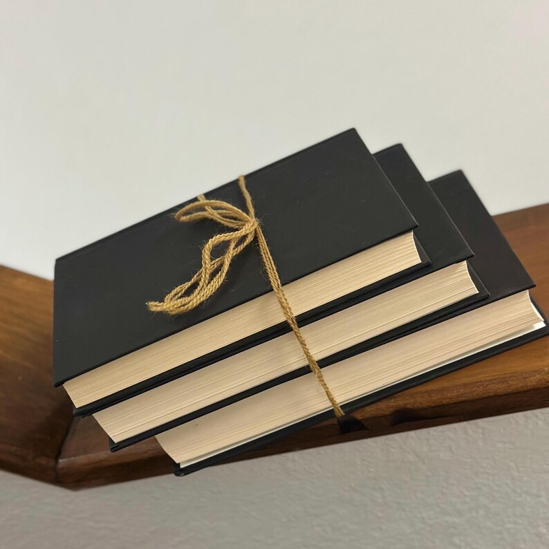 May include: Three black hardcover books tied together with twine. The books have cream-colored pages and are stacked on a wooden shelf. The twine is tied in a decorative bow.