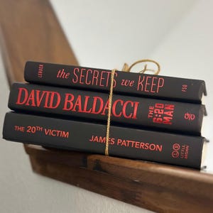May include: Three hardcover books stacked and tied with twine. The books are black with red lettering. Titles include "The Secrets We Keep", "David Baldacci", and "The 20th Victim". The books are on a wooden shelf.