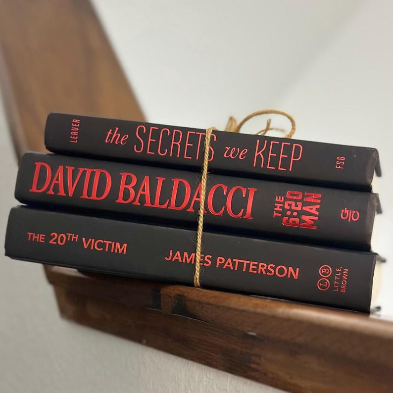 May include: Three hardcover books stacked and tied with twine. The books are black with red lettering. Titles include "The Secrets We Keep", "David Baldacci", and "The 20th Victim". The books are on a wooden shelf.