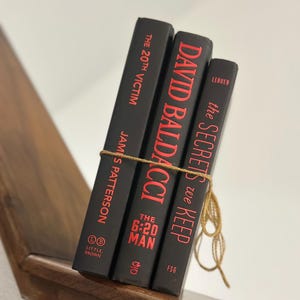 May include: Three hardcover books with black covers and red lettering are tied together with twine. The books are titled "The 20th Victim" by James Patterson, "David Baldacci", and "The Secrets We Keep". The books are on a wooden shelf.