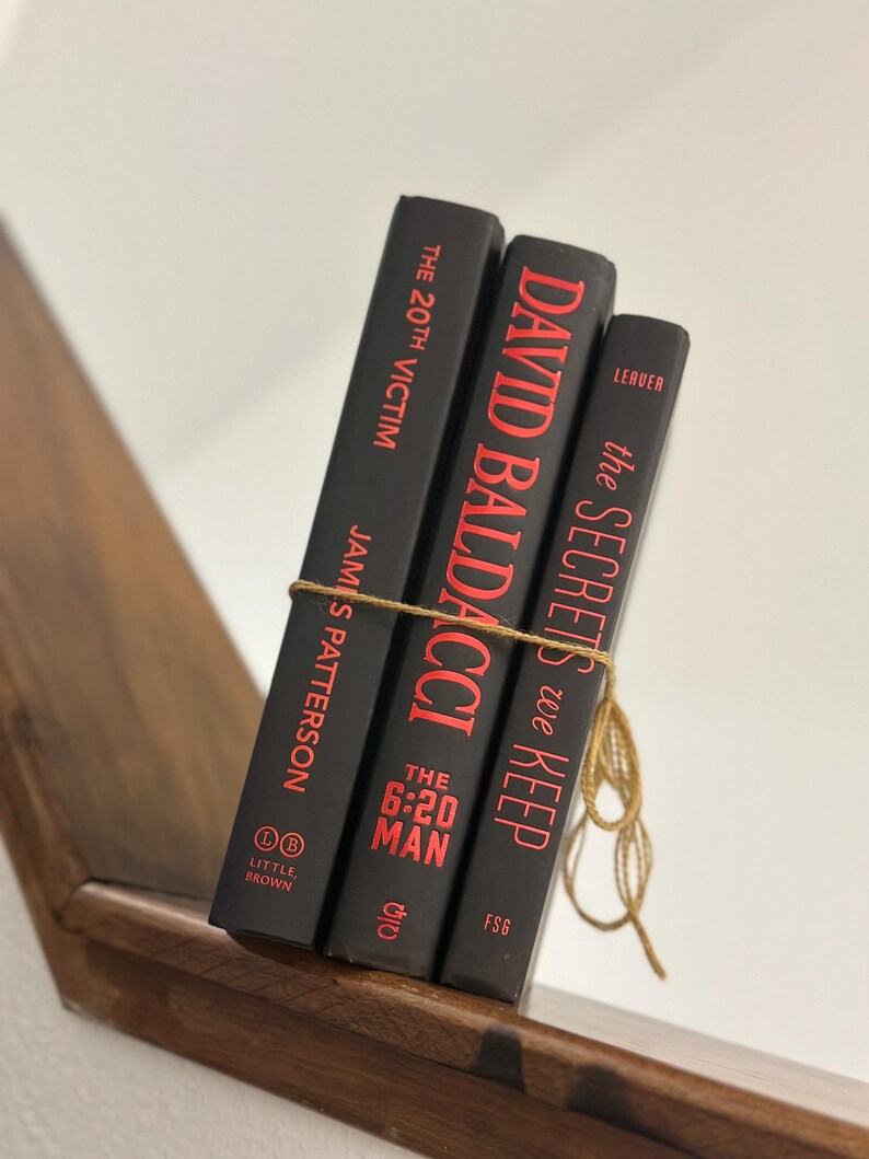 May include: Three hardcover books with black covers and red lettering are tied together with twine. The books are titled "The 20th Victim" by James Patterson, "David Baldacci", and "The Secrets We Keep". The books are on a wooden shelf.