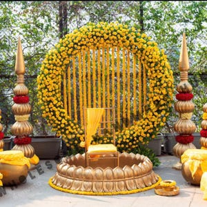 Può includere: Allestimento dorato per cerimonia nuziale con un grande arco floreale circolare di fiori gialli. La scena comprende pilastri decorativi, una sedia dorata e una base a forma di loto, il tutto adornato con accenti gialli e rossi. Il tema generale è festoso.