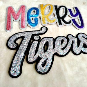 Puede incluir: Parches bordados con detalles de lentejuelas. La palabra "MERRY" está escrita con lentejuelas rosas, azules, amarillas, negras y moradas. La palabra "Tigers" está escrita con lentejuelas plateadas con un borde negro. Parches sobre una superficie blanca texturizada.