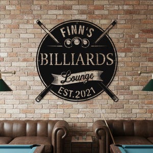 May include: Black metal sign for a billiards lounge, with the text "Finn's Billiards Lounge EST. 2021" in a circular design, crossed by pool cues. The sign is mounted on a brick wall.