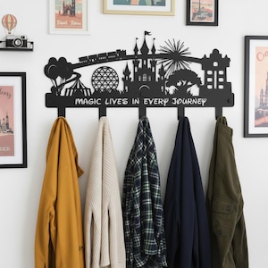 Könnte beinhalten: Schwarzer Wand-Kleiderhaken aus Metall mit einer Silhouette eines Schlosses, Zuges und anderer ikonischer Formen. Der Haken hat vier Haken und den Text "MAGIC LIVES IN EVERY JOURNEY". Verschiedene Mäntel und Jacken hängen an den Haken.