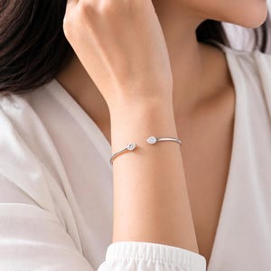 Könnte beinhalten: Ein silberfarbenes, offenes Armreif-Armband mit zwei klaren, funkelnden Edelsteinen. Einer ist rund, der andere birnenförmig. Das Armband wird am Handgelenk getragen und unterstreicht sein minimalistisches Design und seinen eleganten Stil.