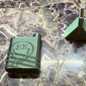 Può includere: Un oggetto rettangolare verde scuro con un design a faccina sorridente e un piccolo oggetto triangolare verde sono esposti su una grande superficie di pietra lucida, viola e bianca. Gli oggetti sembrano essere di metallo.