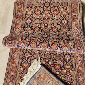 May include: An intricately patterned rug with a navy blue base, featuring floral motifs in red, orange, and green. The rug is partially rolled, revealing a detailed border and fringed edge, showcasing a traditional design.