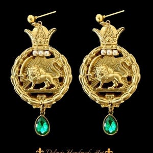 Puede incluir: Un par de pendientes dorados con un emblema de león, una corona de laurel y un diseño de corona. Cada pendiente presenta un colgante de piedra preciosa verde esmeralda en forma de lágrima. Los pendientes están sobre un fondo negro.