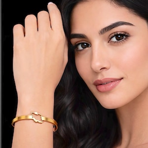 May include: A gold-colored bangle bracelet featuring a cutout design of a country's map. The bracelet is worn on a wrist, with a black background. The bracelet is a simple, elegant piece of jewelry.