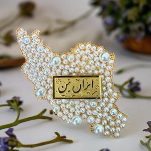Puede incluir: Broche dorada con la forma de Irán, incrustada con perlas blancas. Una placa rectangular dorada con escritura persa está centrada en la broche. La broche está rodeada de flores moradas y tallos verdes.