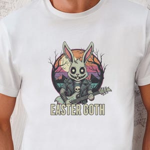 Puede incluir: Camiseta blanca con un gráfico de un conejito con cara de calavera, que lleva una chaqueta de cuero y toca la guitarra. El texto "EASTER GOTH" está debajo del gráfico del conejito. La camiseta la lleva una persona.