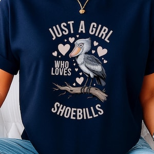 Op de afbeelding: Marineblauwe t-shirt met de tekst "JUST A GIRL WHO LOVES SHOEBILLS". De afbeelding toont een schoenbekooievaar op een tak, omringd door harten. Het shirt is een casual kledingstuk.