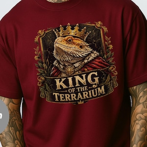 Op de afbeelding: Een bordeauxrood t-shirt met een grafische print van een baardagame die een kroon en een rode cape draagt. De hagedis is omlijst door een decoratieve gouden rand met bloemmotieven. Onder de hagedis staat op een banner de tekst KING OF THE TERRARIUM.