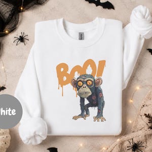 Könnte beinhalten: Ein weißes Langarm-Sweatshirt mit einer Grafik eines zombieartigen Affen mit orangefarbenen Augen und einem abgenutzten Aussehen. Hinter dem Affen ist das Wort BOO in einer tropfenden orangefarbenen Schriftart aufgedruckt. Rundhalsausschnitt.