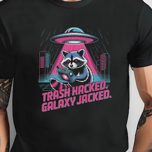 Può includere: T-shirt nero con un disegno di procione all'interno di un UFO, con la scritta "TRASH HACKED. GALAXY JACKED." Il procione tiene in mano un dispositivo, con uno skyline sullo sfondo. Il design utilizza colori rosa, blu e verde.