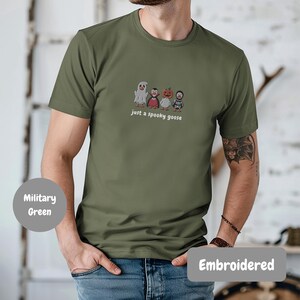 A man wearing a green t-shirt with a graphic of four cartoon characters, including a ghost, a pumpkin, a witch, and a goose, along with the text "Just a spooky goose".