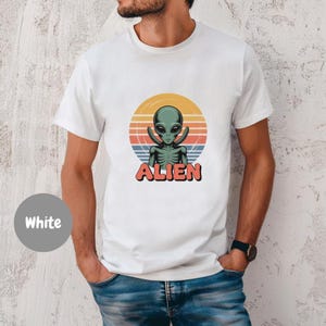 Puede incluir: Camiseta blanca con un diseño retro de un alienígena verde y la palabra "ALIEN" en rojo. El alienígena está situado sobre un círculo con los colores del atardecer. Prenda de vestir informal.