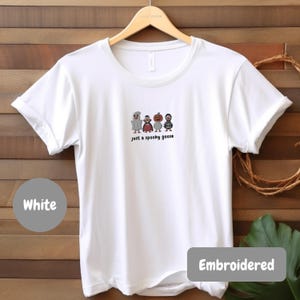A white t-shirt with a graphic design of four cartoon characters and the text "Just a spooky goose" printed on it, hanging on a wooden hanger.