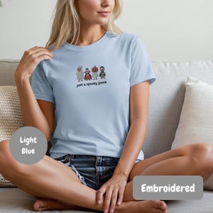 A woman wearing a light blue t-shirt with a graphic of a spooky goose and the text "Just a spooky goose" is sitting on a couch.