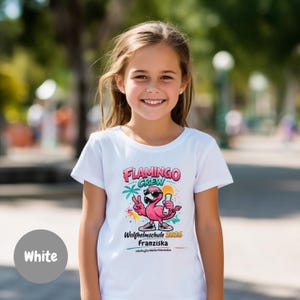 Könnte beinhalten: Weißes T-Shirt mit einem bunten Aufdruck eines Flamingos mit Sonnenbrille und Getränk. Der Text "FLAMINGO CREW" steht über dem Flamingo, darunter "Wolfhelmschule 2026 Franziska". Das T-Shirt wird von einem lächelnden Kind getragen.