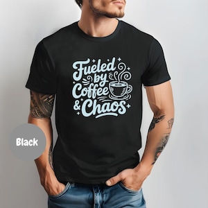 Può includere: T-shirt nero con la scritta "Fueled by Coffee & Chaos" in azzurro chiaro, con un carattere stravagante. Il design include una tazza di caffè. La maglietta è a girocollo.