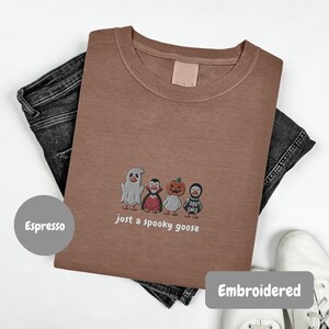 A brown t-shirt with a Halloween-themed embroidered design, featuring four cartoon characters, including a ghost, skeleton, pumpkin, and goose, along with the text "just a spooky goose".