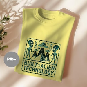 Puede incluir: Camiseta amarilla clara con un diseño verde azulado. El diseño incluye extraterrestres, pirámides y platillos volantes. El texto dice "BUILT WITH ALIEN TECHNOLOGY".